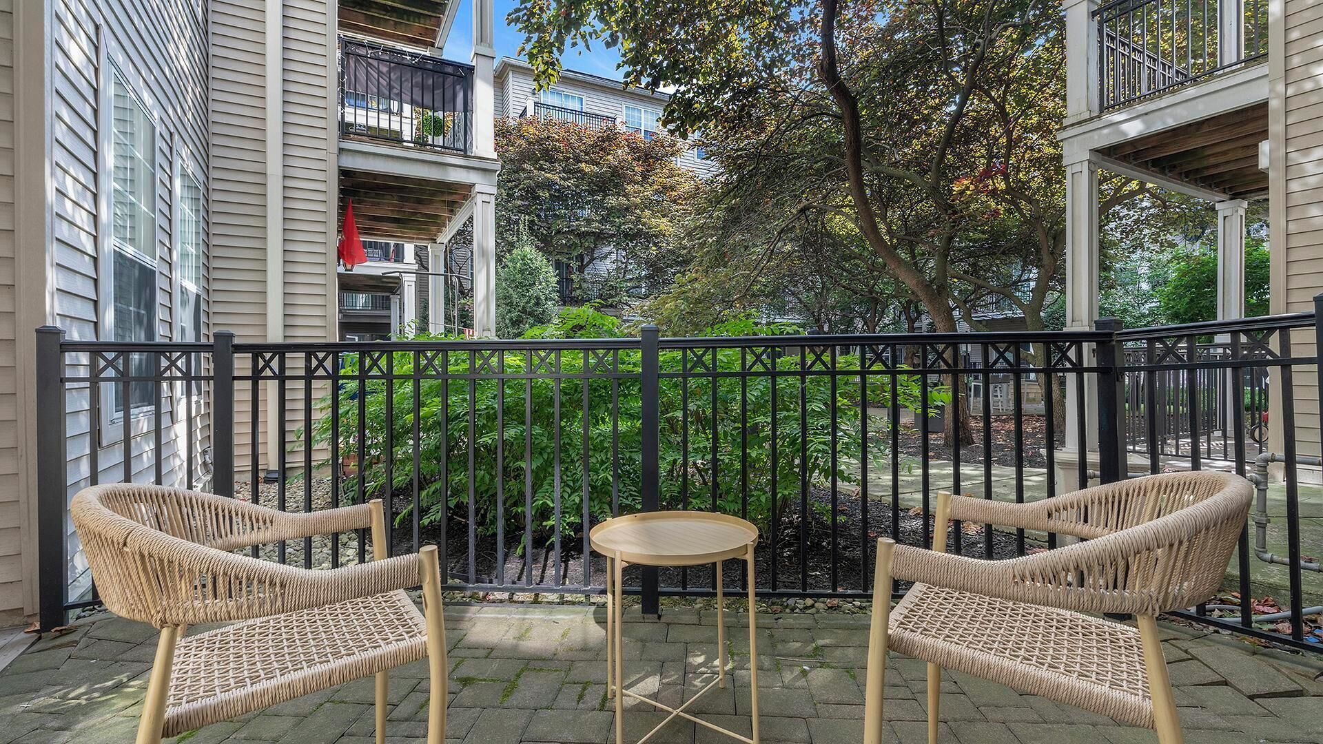Ein Balkon mit Korbstühlen und einem kleinen Tisch, umgeben von einem schwarzen Metallzaun, mit Blick auf einen Garten mit Bäumen und einem Gebäude im Hintergrund.