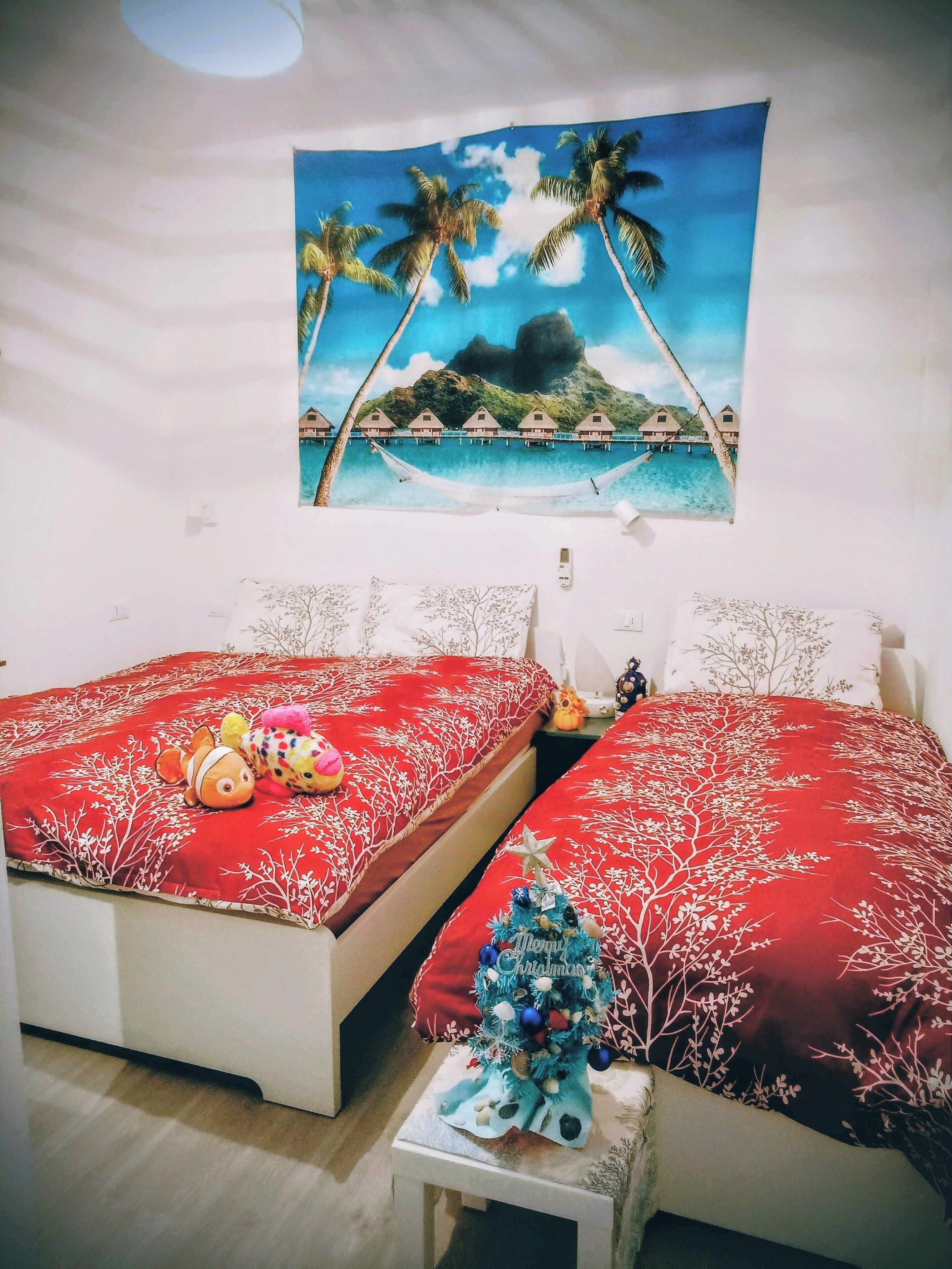 A bedroom with two beds, each with a red bedspread and white pillows. There is a wall hanging above the beds depicting a tropical scene with palm trees and a beach.