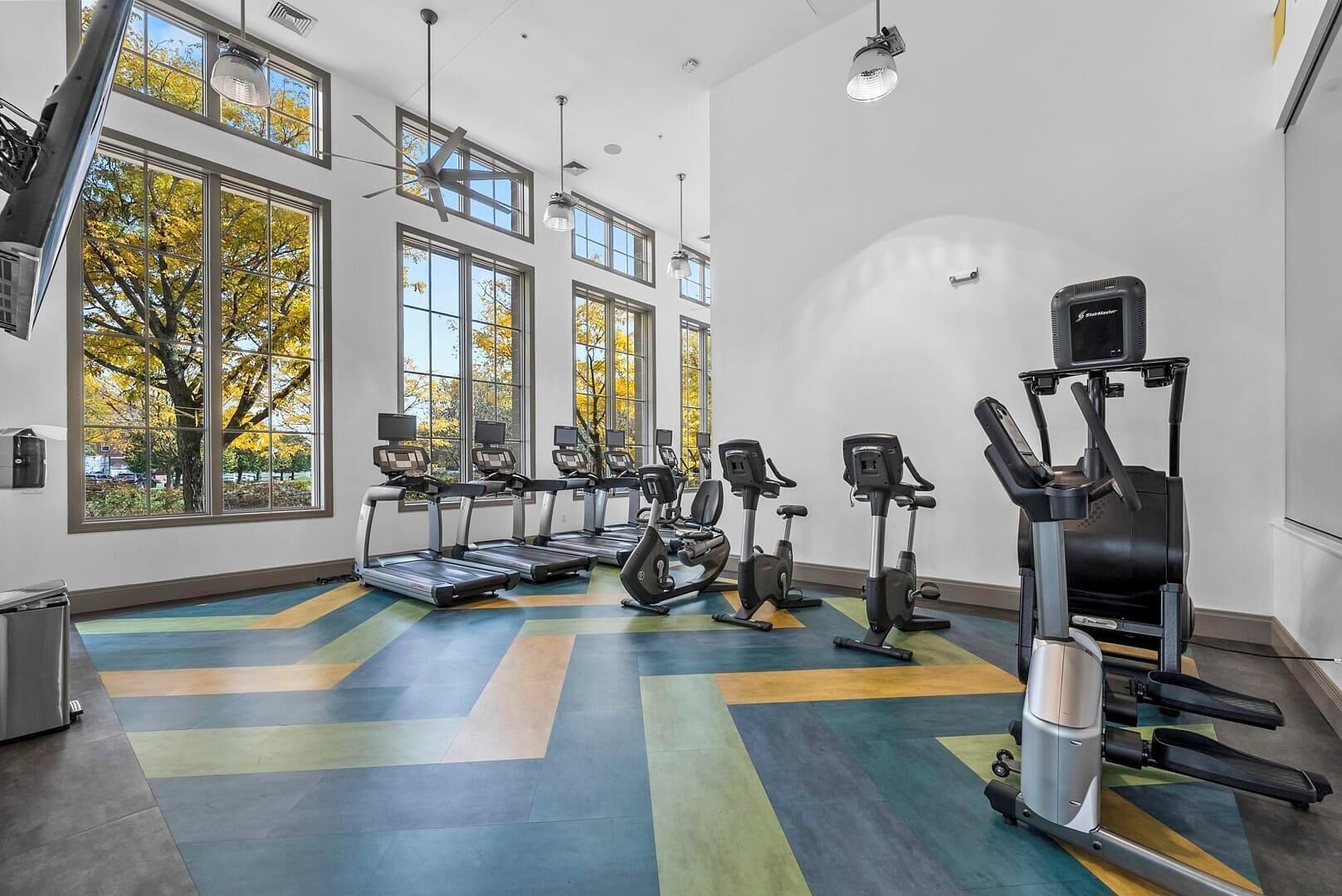 A gym with treadmills, elliptical machines, and a stationary bike, large windows, and a colorful floor.