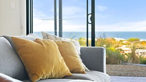 Un canapé avec deux coussins, une fenêtre donnant sur la mer et la verdure, et une structure métallique en arrière-plan.