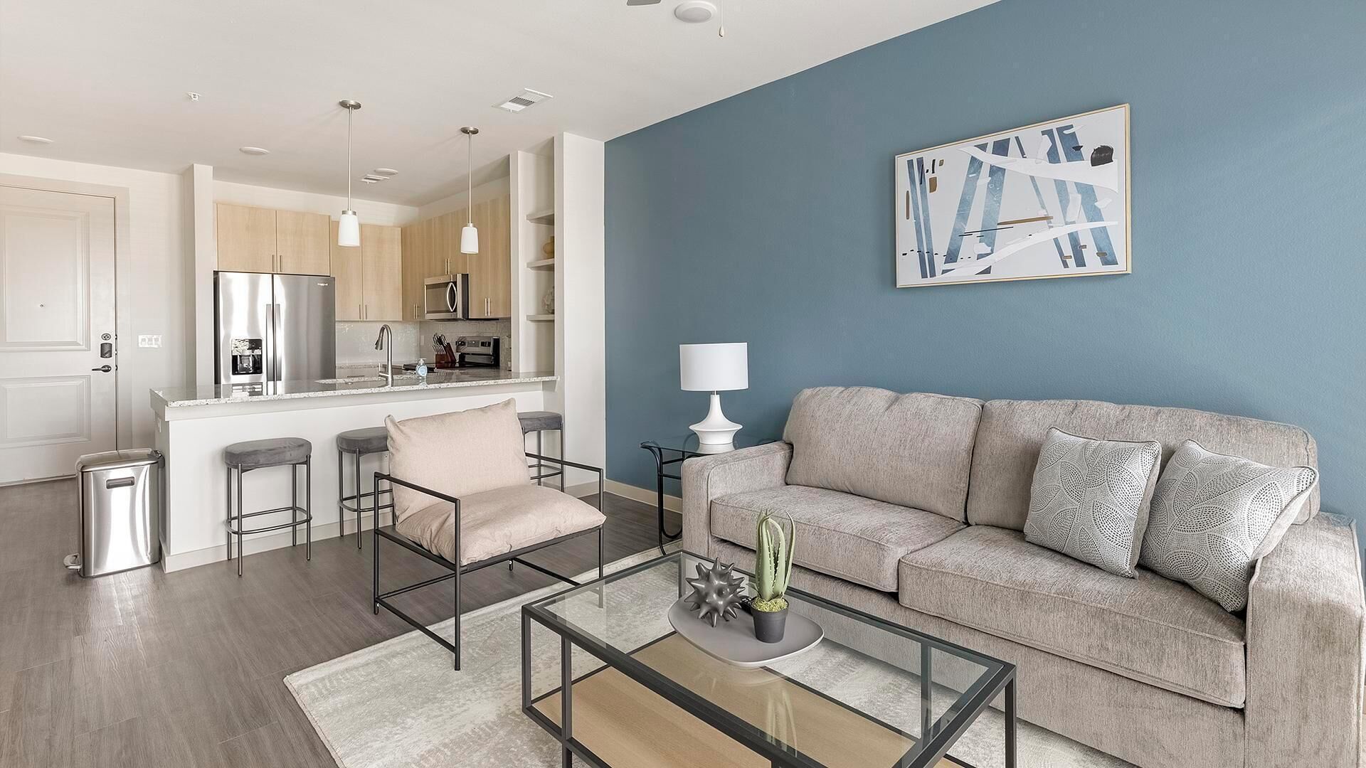 A modern living room with a sofa, chairs, a glass coffee table, and a kitchen in the background.