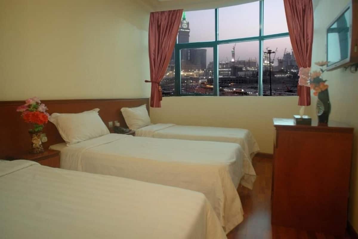 Ein Hotelzimmer mit zwei Betten, einem Nachttisch mit Blumenvase, einem Holztisch und einem Fenster mit Blick auf die Stadt.