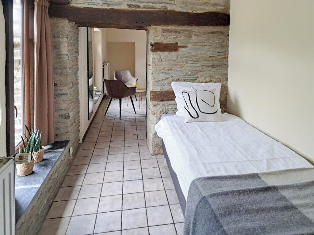 A narrow hallway with a bed on the right, a chair in the background, and a stone wall.