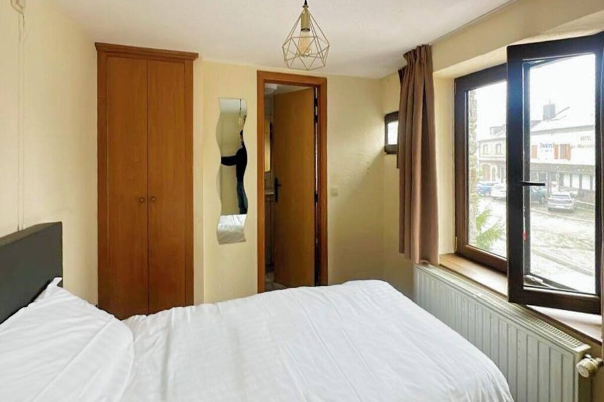 A bedroom with a bed, a wooden wardrobe, a mirror, a window with curtains, and a radiator.