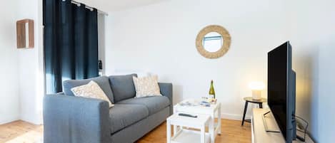Ett modernt vardagsrum med en grå soffa, ett vitt soffbord, en tv och ett litet sidobord med en flaska och en fjärrkontroll.