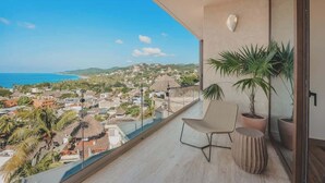 Een balkon met helder uitzicht op zee en een stad. Er staat een stoel en een plant in pot op het balkon.