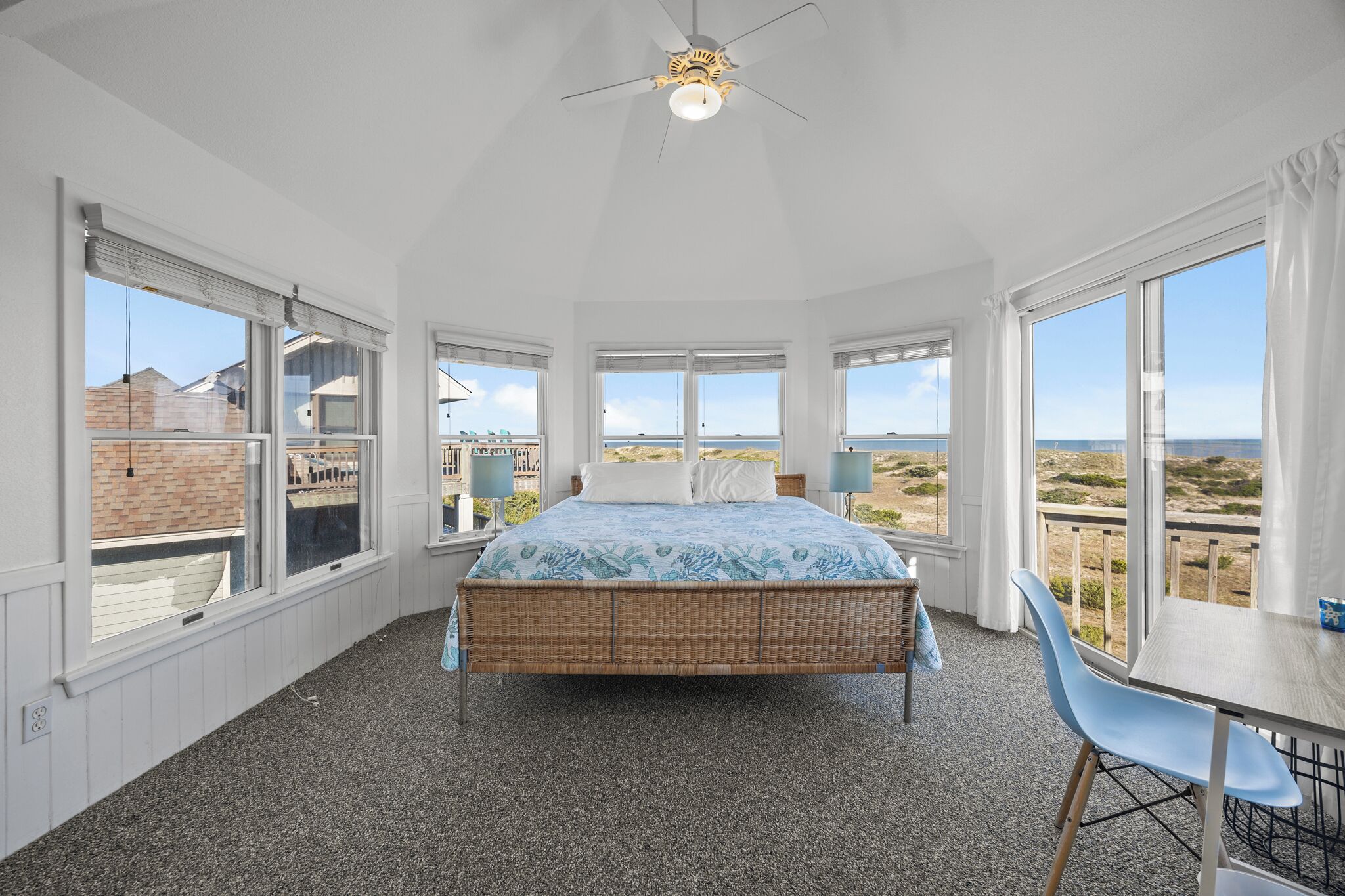 A bedroom with a bed, a table, and chairs, large windows with a view of the beach and buildings.