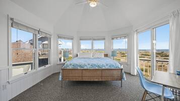 A bedroom with a bed, a table, and chairs, large windows with a view of the beach and buildings.