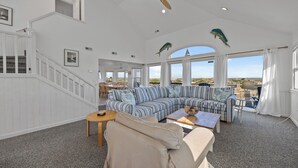 A spacious living room with a large striped sofa, a beige armchair, and a wooden coffee table. There is a staircase with white railings and a ceiling fan. - Moon Palace: Oceanfront | Elevator | Hot Tub | Private Pool | Game Room (Salvo)