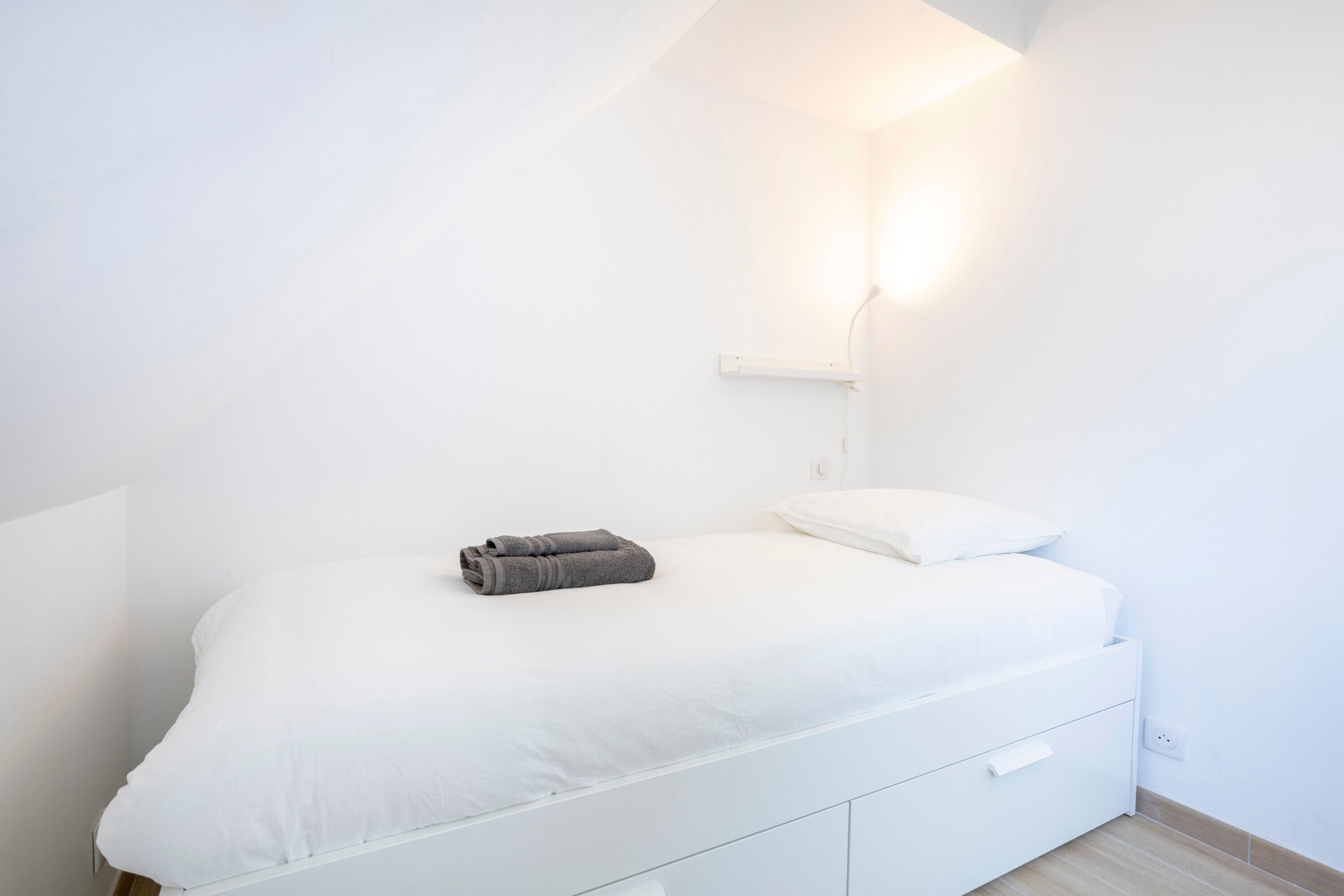 Een wit bed met een kussen en een opgerolde handdoek, een nachtkastje en een wandlamp.