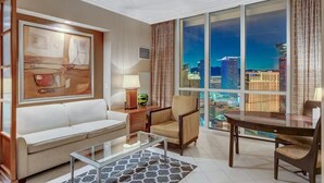 A modern living room with a large window offering a city view, a white sofa, a glass coffee table, and a wooden side table with a lamp. - F1 View + Strip Views, 34th Floor at MGM Signature (Las Vegas)