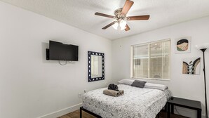 Ein Schlafzimmer mit einem Bett, einem Deckenventilator, einem Fernseher, einem Fenster mit Jalousien und Wandbildern.