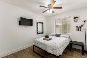 Un dormitorio con cama, ventilador de techo, televisión, ventana con persianas y cuadros en la pared.