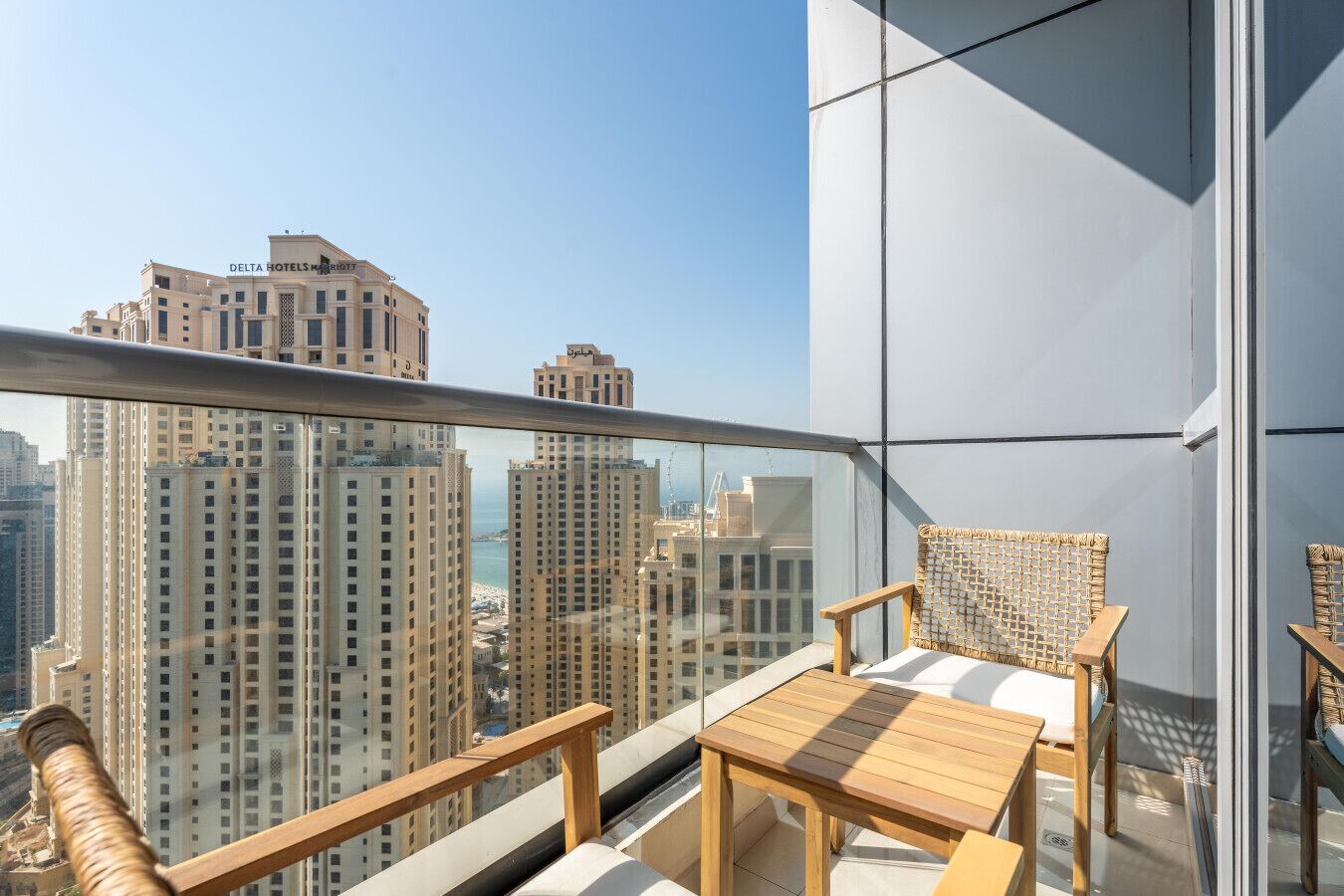 A balcony with a view of high-rise buildings and the sea. There are two wicker chairs and a small wooden table.