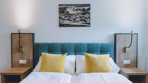 Una cama doble con ropa blanca, dos almohadas amarillas y un cabecero verde azulado. Hay dos mesitas de noche de madera con lámparas y un enchufe en la pared. Sobre la cama, se exhibe una fotografía blanco y negro de un paisaje montañoso nevado.
