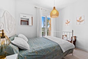 A bedroom with a bed, a hanging light fixture, a window with curtains, and a view of palm trees.
