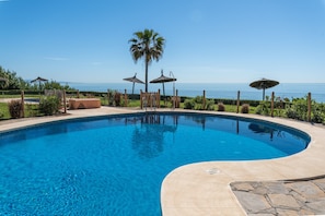 A swimming pool with a curved edge, surrounded by a fence, palm trees, and umbrellas.