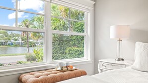 Ein Schlafzimmer mit großem Fenster, einem Nachttisch mit Lampe, einem Bett mit weißer Bettdecke und einer kleinen Bank mit Tablett mit Essen.