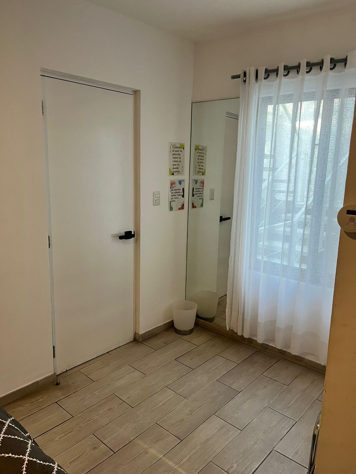 A room with a wooden floor, a white door, a mirror, and a window with sheer curtains.