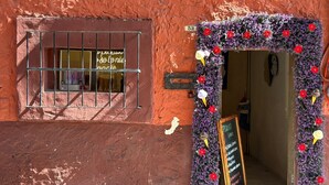 A building entrance with a window displaying a sign in a foreign language, a door decorated with purple flowers and red poinsettias, and a chalkboard menu outside. - NIDO Hotel (San Miguel de Allende)