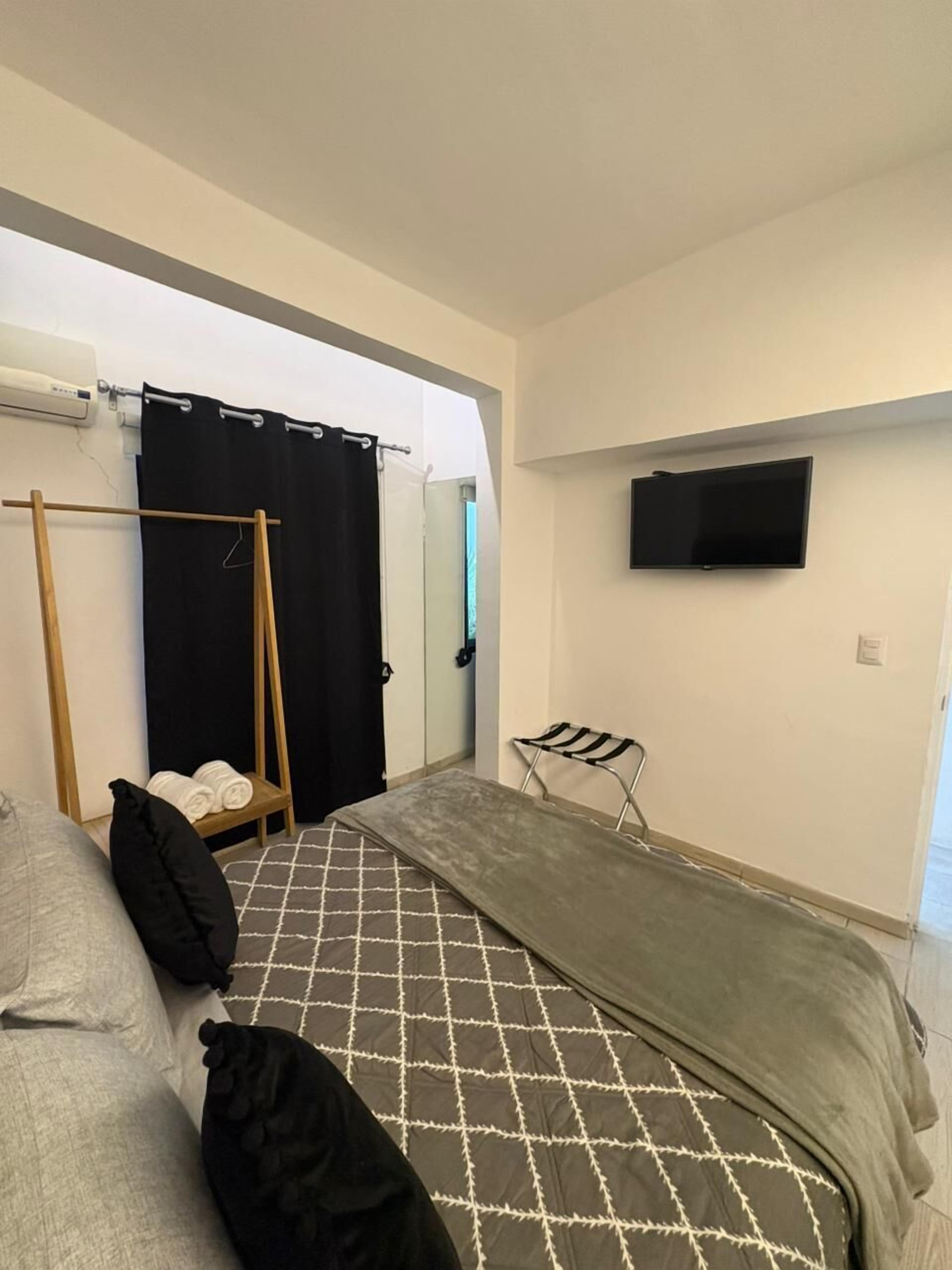 Una habitación pequeña con cama, una mesita con toallas, una silla, televisor montado en la pared y una ventana con cortinas.