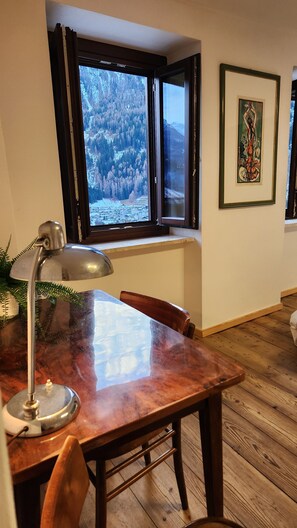 Een eethoek met een houten tafel, stoelen, een bureaulamp, een raam met uitzicht op een bos en een ingelijst schilderij aan de muur.