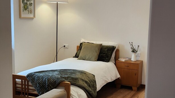 Een slaapkamer met een bed, nachtkastje, lamp en een stoel.