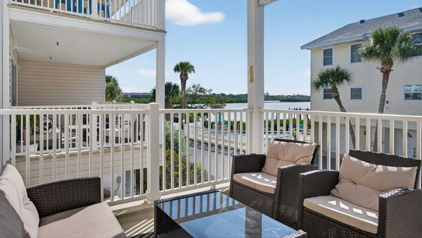 A covered outdoor area with wicker furniture and a glass table, overlooking a pool and buildings. - 🌅 Captain’s Cove 402 | 2BR Condo Across from Indian Shores Beach (Indian Shores)