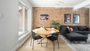 A modern living room with a brick wall, a round wooden dining table, black chairs, a black sofa, and a large window. - MTL Central Stay Apartments (Montréal)