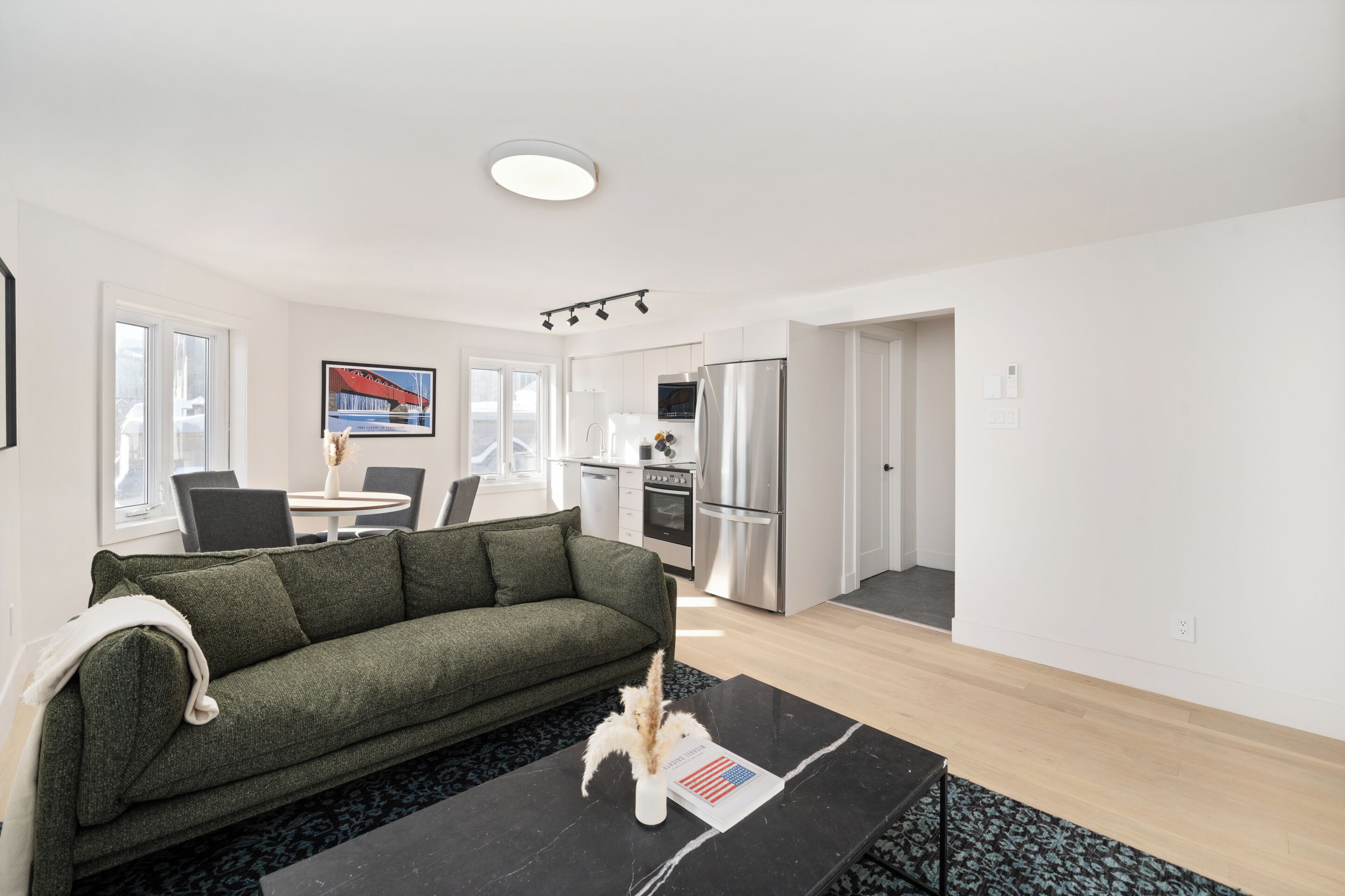 A modern living room with a sofa, a coffee table with a vase and a flag, a dining area with chairs and a table, and a kitchen with appliances.