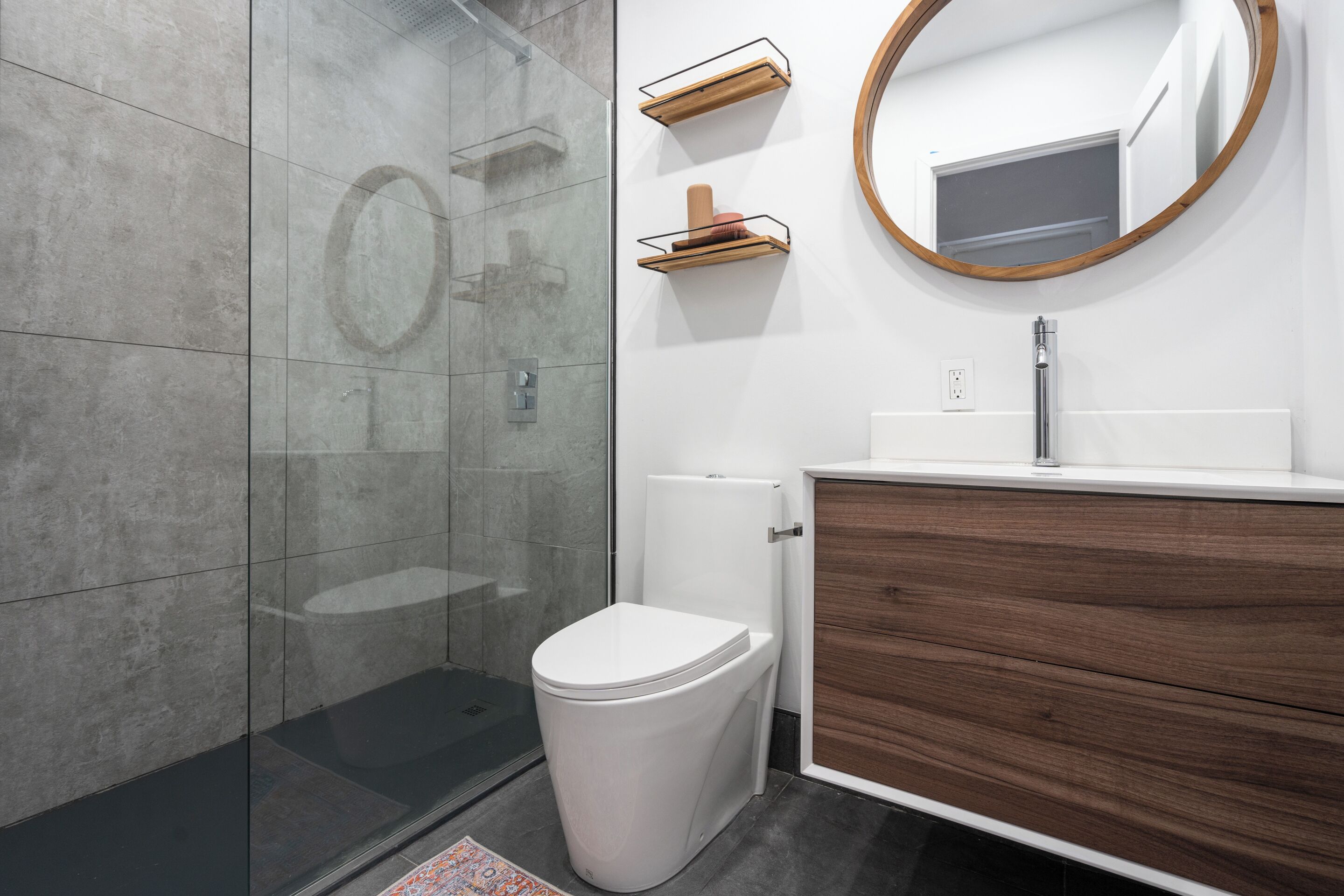 A modern bathroom with a glass shower enclosure, a white toilet, a wooden vanity with a mirror, and built-in shelves.