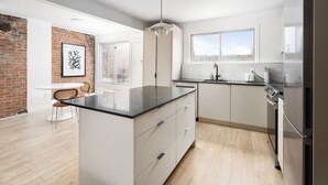 A modern kitchen with a central island, white cabinetry, a hanging light fixture, and a brick accent wall. - MTL Central Stay Apartments (Montréal)