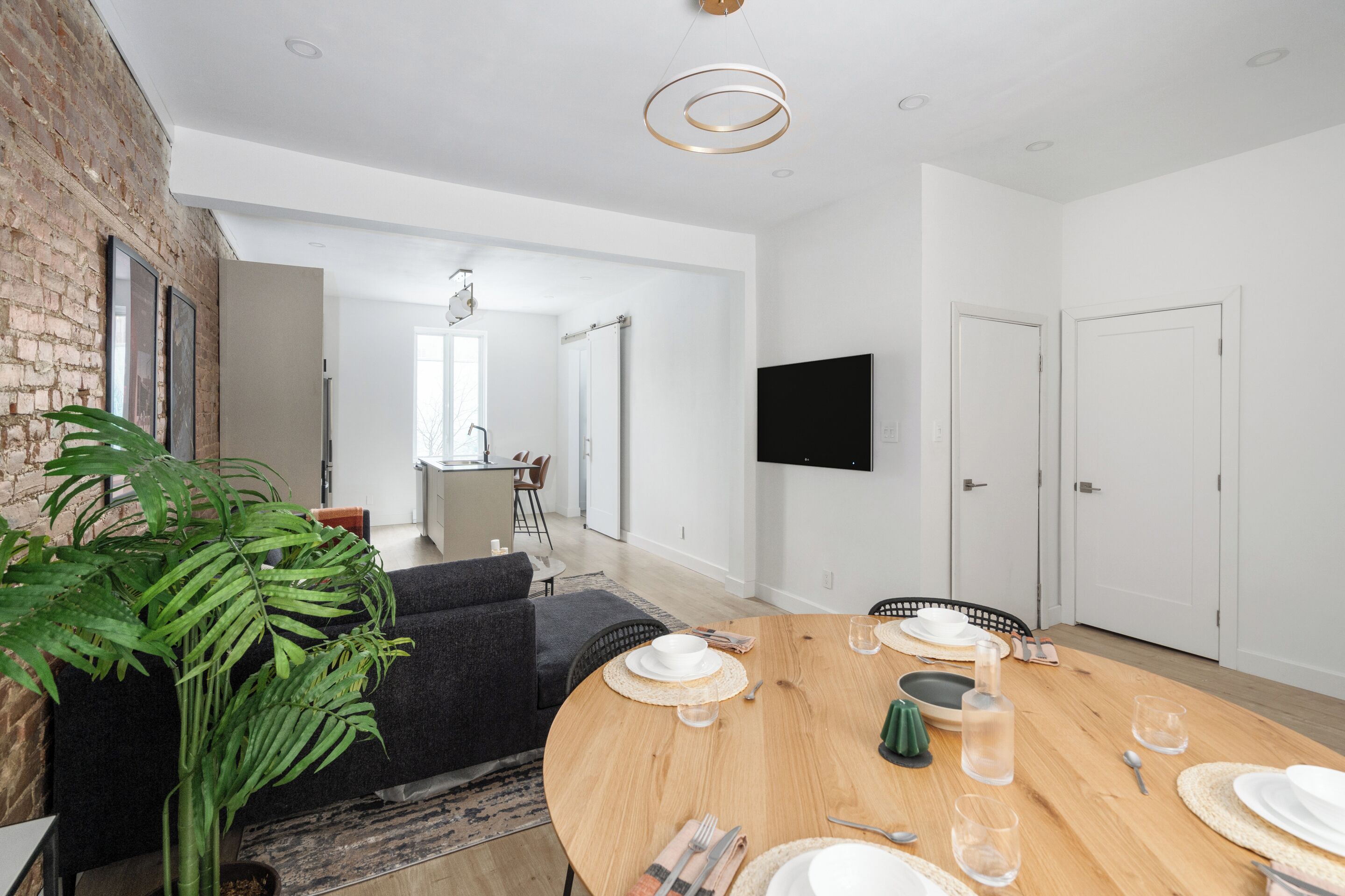 Un espace repas moderne avec une table ronde en bois, dressée pour quatre personnes, une grande plante en pot et un canapé gris.