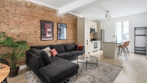 A modern living room with a brick wall, a dark grey sofa, a small coffee table, and a potted plant. - MTL Central Stay Apartments (Montréal)