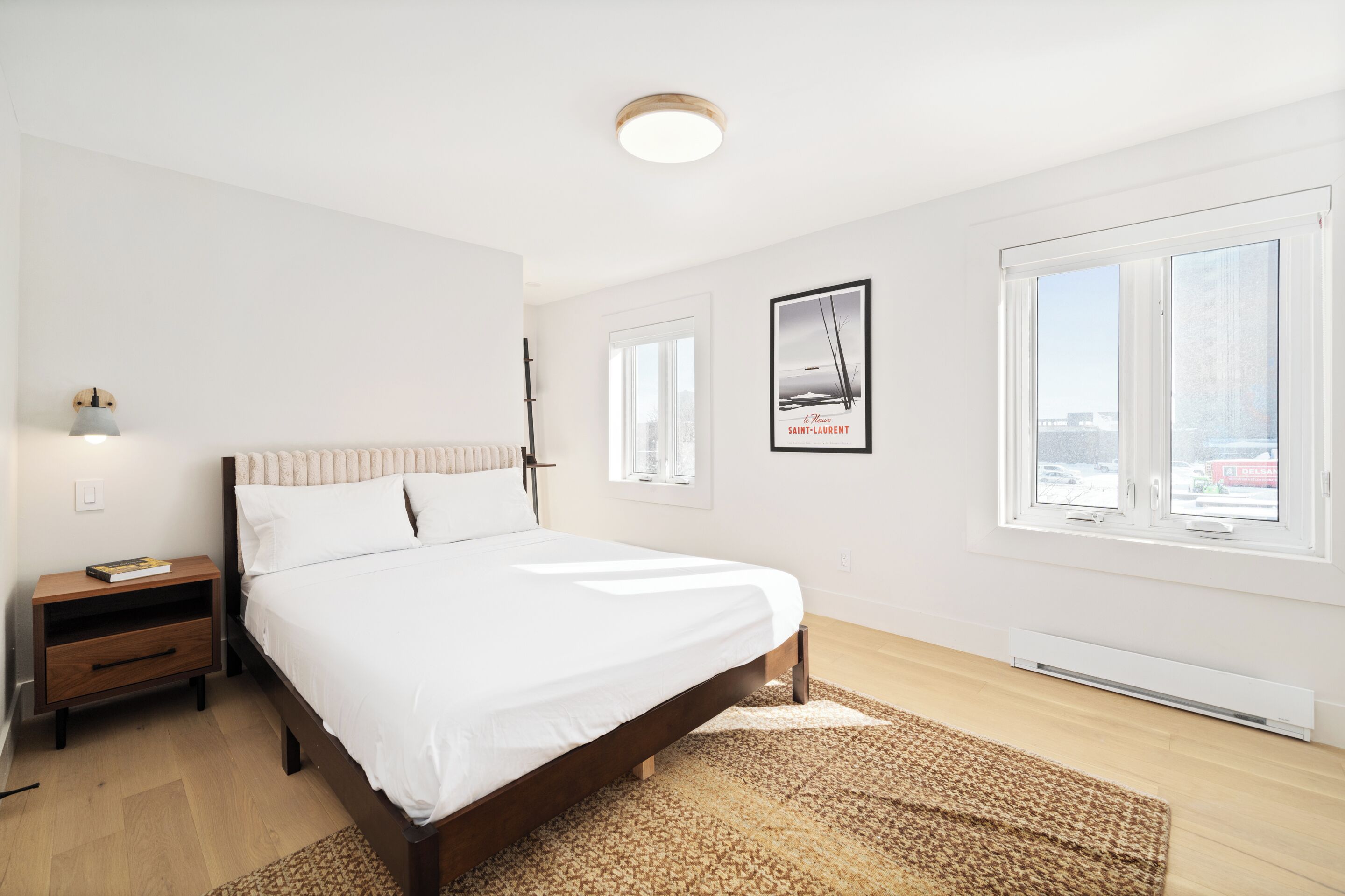 Een slaapkamer met een bed, nachtkastje, raam en een lijst met een afbeelding aan de muur.
