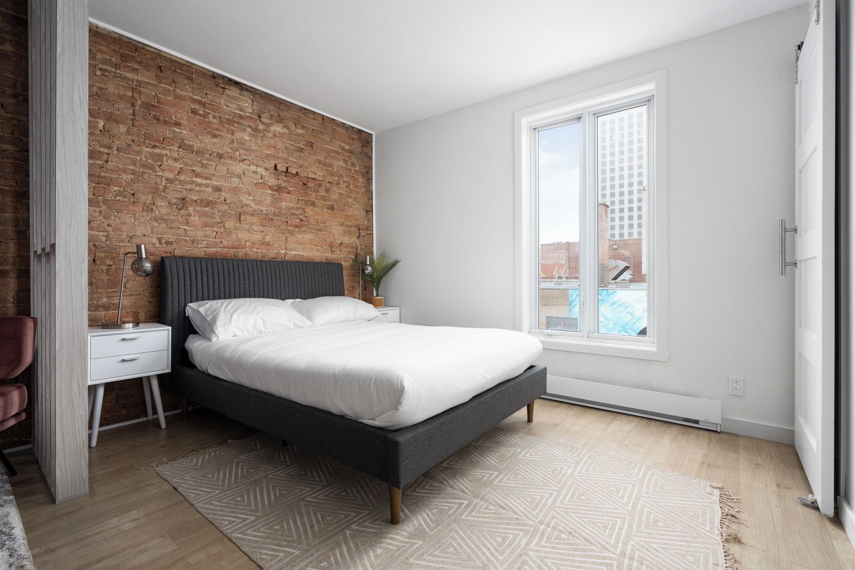 Een moderne slaapkamer met een bakstenen muur, een groot raam, een bed, een nachtkastje en een stoel.