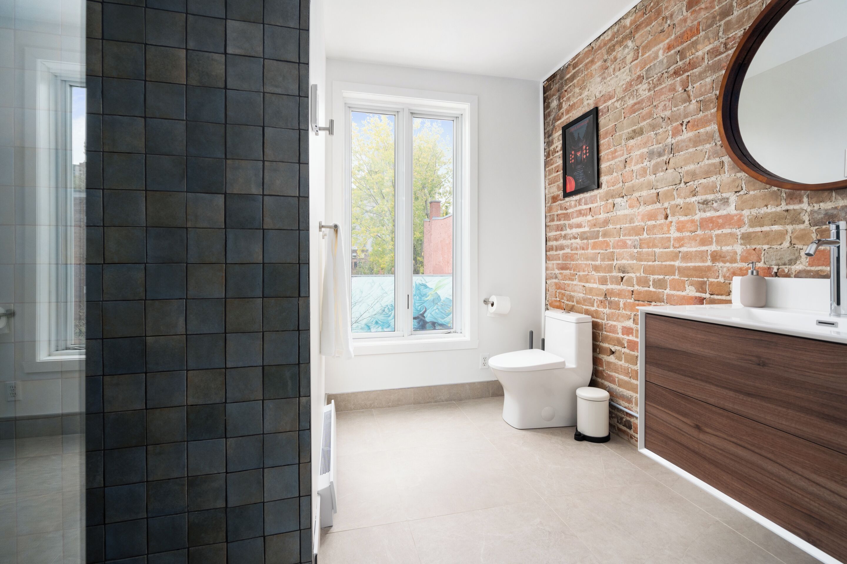 Un baño moderno con pared de ladrillo, inodoro blanco, lavamanos con espejo y ventana.