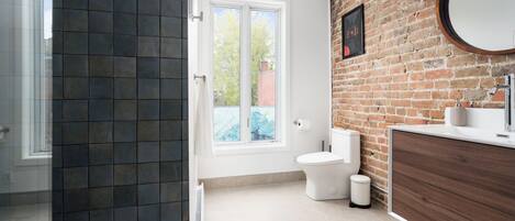 Un baño moderno con pared de ladrillo, inodoro blanco, lavamanos con espejo y ventana.