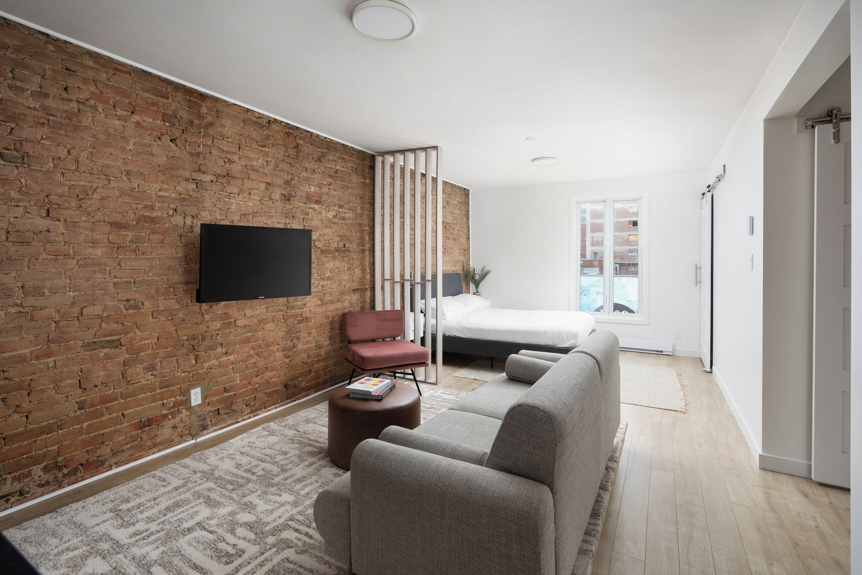 Un salon moderne avec un mur en briques, un téléviseur à écran plat, un canapé, un fauteuil et un lit à l’arrière-plan.