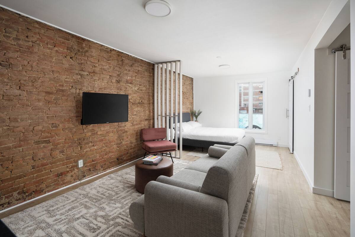 A modern living room with a brick wall, a flat-screen TV, a sofa, a chair, and a bed in the background.