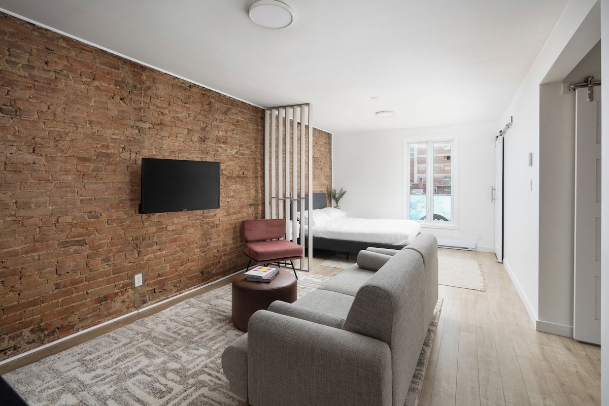 A modern living room with a brick wall, a flat-screen TV, a sofa, a chair, and a bed in the background.