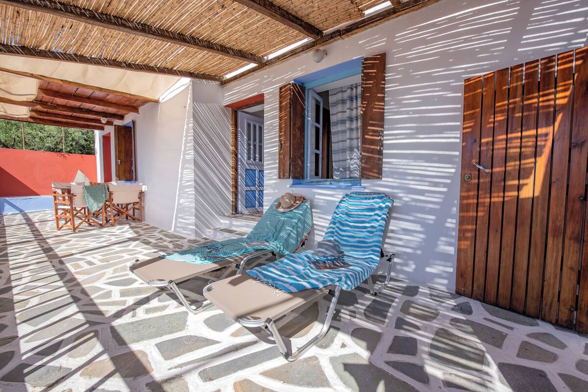 Un'area relax esterna coperta, con poltrone e un tavolo, una porta in legno e una finestra con persiane.
