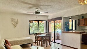 Ein gemütlicher Wohnbereich mit einer Couch, einem kleinen runden Tisch und einem Esstisch mit Stühlen. Ein Deckenventilator, eine Küchenzeile mit Arbeitsplatte und ein großes Fenster mit Blick ins Freie sind ebenfalls vorhanden.