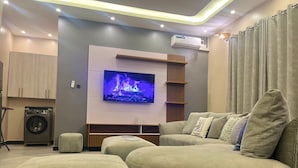En moderne stue med et fladskærms-tv, en sofa, skottomanner og en vaskemaskine, samt en loftslampe.
