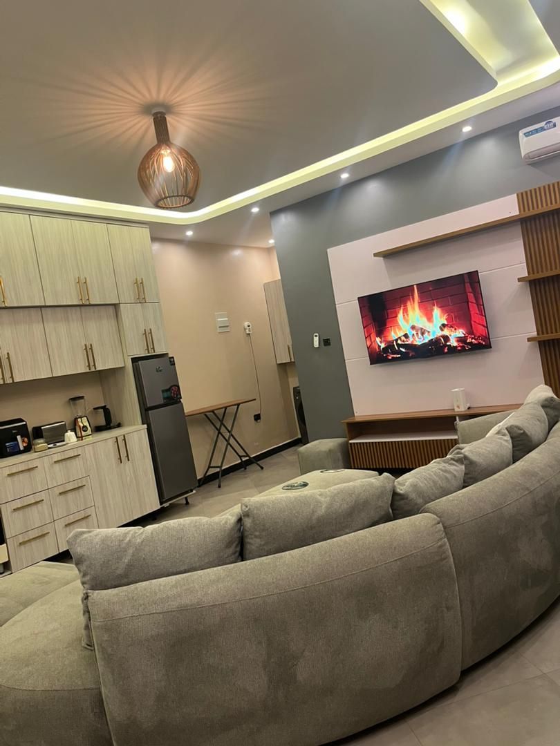 Een moderne woonkamer met een grote flatscreen televisie aan de muur, een beige bank en een keukenhoek met kasten en apparatuur.