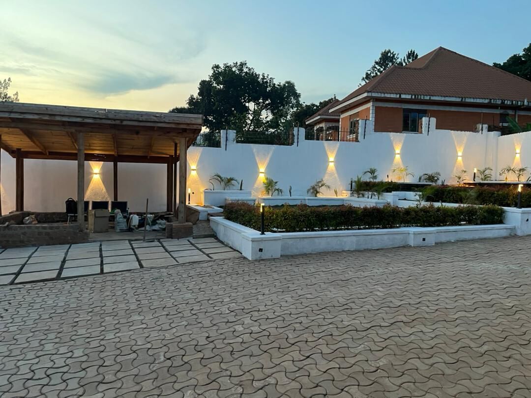 A covered outdoor area with a tiled floor, a white wall with embedded lighting, and a building with a brown roof in the background.