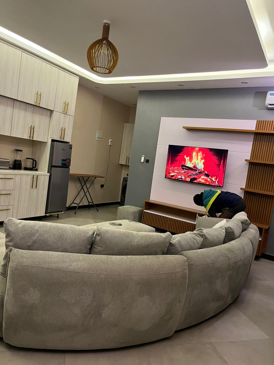 Una sala moderna con un amplio sofá en forma de L, un televisor de pantalla plana montado en una pared de madera, una zona de cocina con armarios de colores claros y una lámpara colgante.