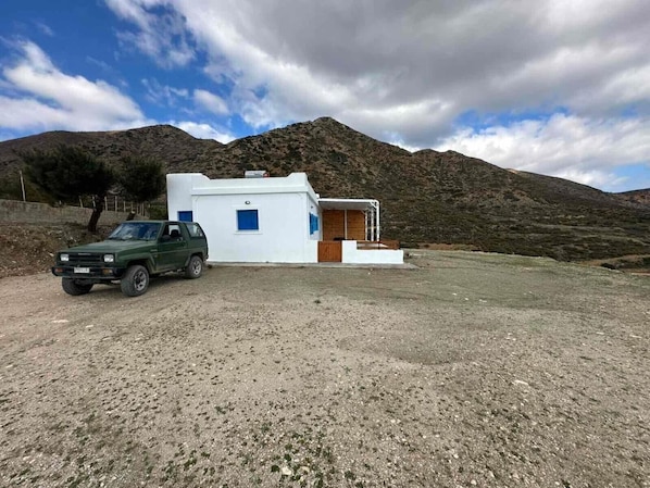 A small, white building with a wooden door and steps leading up to it, situated in a rural area with a mountain in the background.