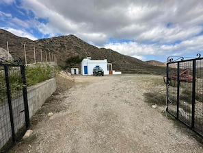 A gravel driveway leading to a white building with a blue door, surrounded by mountains and a cloudy sky.