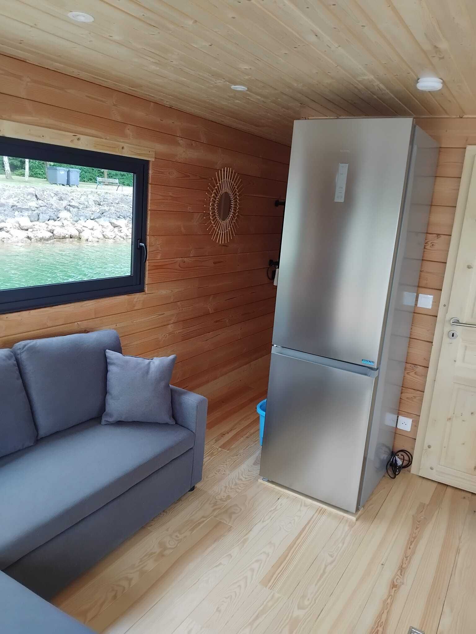 Un espace de vie compact comprenant un canapé gris, un réfrigérateur en acier inoxydable, des murs en bois et une fenêtre donnant sur un espace extérieur rocheux.